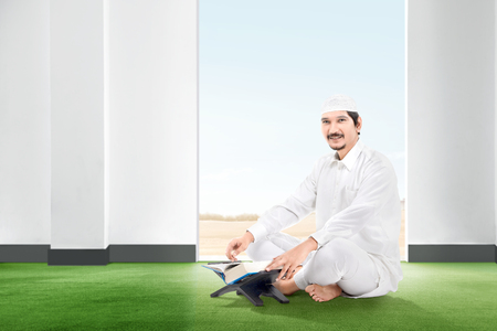 Smiling asian muslim man sitting on the carpet with koran inside the room with open door and blue sky backgroundの写真素材