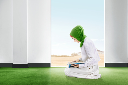 Young asian woman in veil sitting on the carpet reading the koran inside the room with open door and blue sky backgroundの写真素材