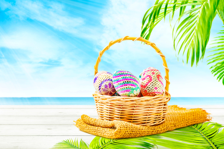 Colorful easter eggs on wooden basket with fabric and palm leaf mat on wooden table with palm branch and blue ocean view over blue sky background. Happy Easterの写真素材
