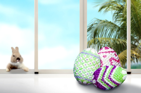 Colorful easter eggs on the table and little bunny on the window glass with palm tree and blue sky background. Happy Easterの写真素材