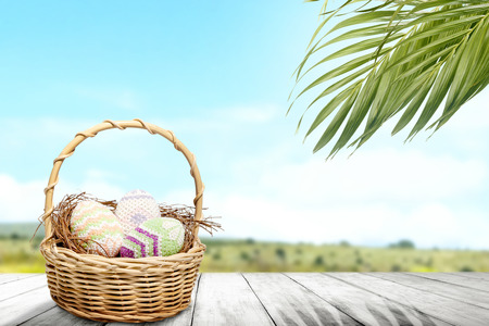 Colorful easter eggs on the nest in wooden basket on wooden table with palm branches and landscape view on blue sky background. Happy Easterの写真素材
