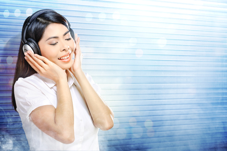 Pretty asian woman in white shirt listening music with headphones over white roller shutter door with smoke and colorful light reflection backgroundの写真素材