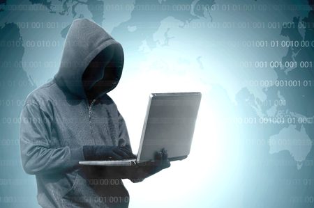 Hacker in black hoodie holding laptop with binary code on virtual screen and world map backgroundの写真素材