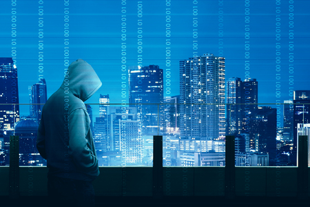 Rear view of hacker in black hoodie standing on the building terrace looking at cityscapes at night with digital binary code backgroundの写真素材