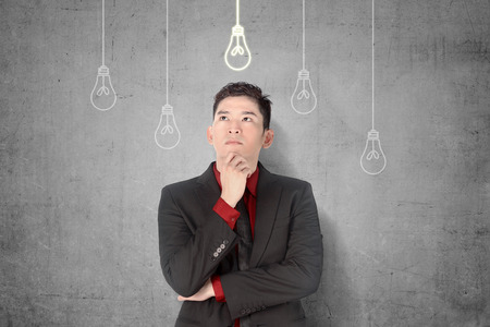 Young asian businessman thinking for new innovative idea with bright light bulbs hanging over gray wall backgroundの写真素材