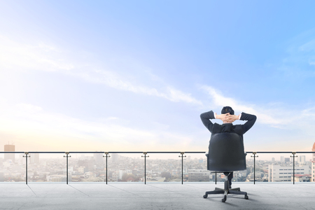 Rear view of asian businessman sitting on the office chair in modern terrace relax and looking at view on the city over blue sky backgroundの写真素材