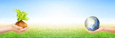 Two human hands holding young plant in the pots and earth planet on the green grass with blue sky background. Earth day conceptの写真素材