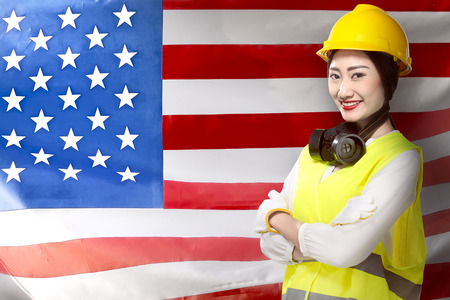 Smiling asian worker woman in safety vest, gloves, yellow helmet and protective mask standing against USA Flag background. Labor day conceptの写真素材
