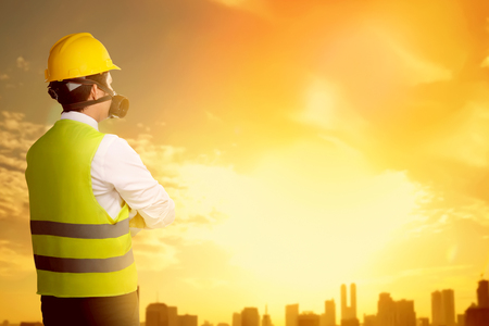Rear view of asian worker man in safety vest, gloves, yellow helmet and protective mask standing and looking at city with sunset backgroundの写真素材