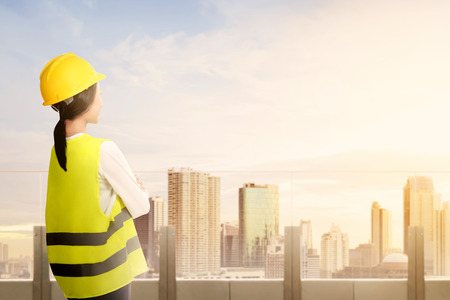Rear view of asian worker woman in safety vest, gloves, yellow helmet and protective mask standing on modern terrace and looking at city with blue sky backgroundの写真素材