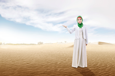 Pretty asian muslim woman with open palm showing something on the sand dune with blue sky backgroundの写真素材