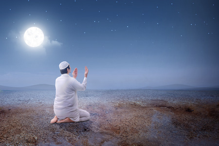 Rear view of asian muslim man sitting in pray position while raised hands and praying on the sand dune with moon and night scene backgroundの写真素材