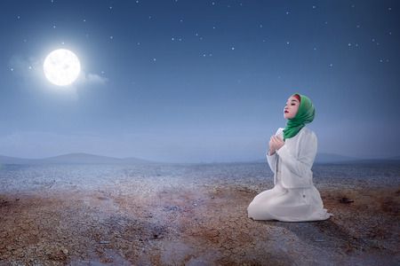 Young asian muslim woman sitting in pray position while raised hands and praying on the sand dune with moon and night scene backgroundの写真素材