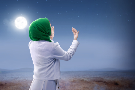Rear view of asian muslim woman in veil raised hands and praying on the sand dune with moon and night scene backgroundの写真素材