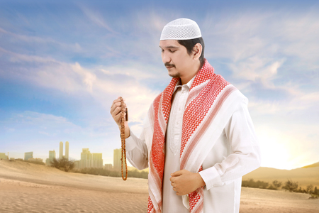Young asian muslim man with cap and turban standing and praying with prayer beads on the sand with blue sky backgroundの写真素材