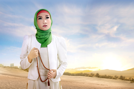 Young asian muslim woman in veil standing and praying with prayer beads on the sand with blue sky backgroundの写真素材