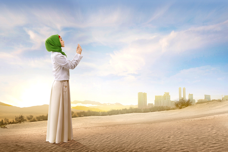 Pretty asian muslim woman in veil standing and praying with raised arms on the sand with blue sky backgroundの写真素材