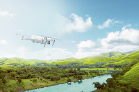 White drone with camera flying over green hills with river and trees over blue sky background