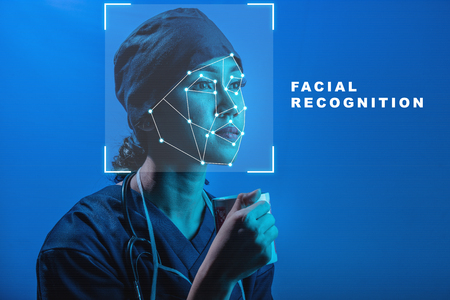 Pretty asian female doctor in surgery uniform and stethoscope holding the glass using face recognition over blue background. Modern technology conceptの写真素材