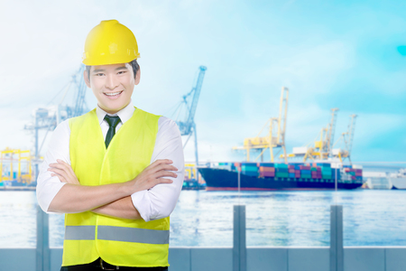 Handsome asian worker with yellow hard hat standing on the office with industrial harbor and blue sky backgroundの写真素材