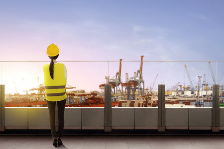 Rear view of asian female worker with safety vest and hard hat standing on office terrace and looking at dock with sunset view backgroundの写真素材