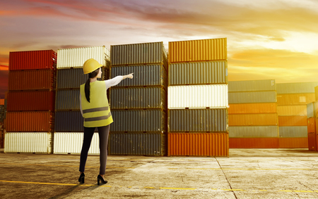 Rear view of asian female worker with safety vest and hard hat standing on the dock and pointing a container with sunset view backgroundの写真素材