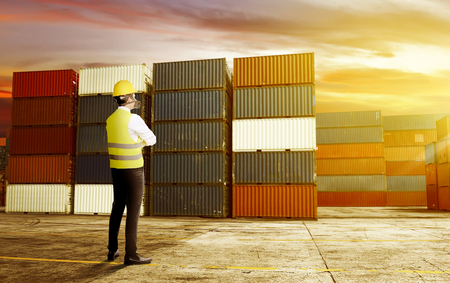 Rear view of asian male worker with safety vest, hard hat and protective mask standing on the dock and looking at pile of container with sunset view backgroundの写真素材