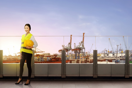 Pretty asian female worker with safety vest holding the hard hat standing on office terrace with dock and sunset view backgroundの写真素材
