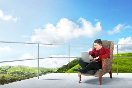 Happy asian businessman sitting on the chair and using laptop on the terrace with green landscapes view and blue sky backgroundの写真素材