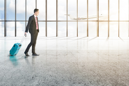 Smiling asian businessman with blue suitcase walking on the airport hall with window of glass and flying airplane over blue sky backgroundの写真素材