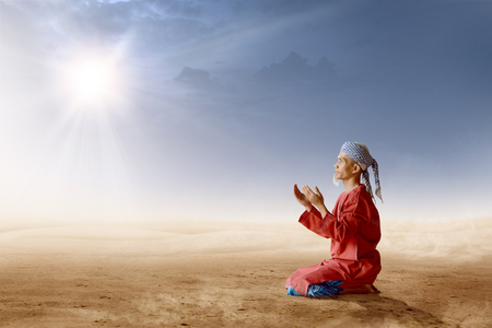 Asian muslim man with turban in his head kneeling and praying with raised hands on desert with sun rays and dark sky backgroundの写真素材