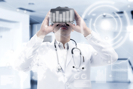 Asian male doctor in white coat and stethoscope looking at digital interface with virtual reality device display the world map, graphic bar and binary code. Augmented reality technologyの写真素材