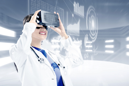 Asian female doctor in white coat and stethoscope looking at digital interface with virtual reality device display the world map, graphic bar and binary code. Augmented reality technologyの写真素材