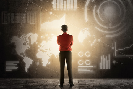 Rear view of asian businessman standing and looking at virtual screen which display the interface of financial chart and world maps. Digital technology conceptの写真素材