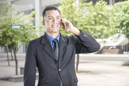 Smiling asian businessman with mobile phone using face recognition over outdoor background. Modern technology conceptの写真素材