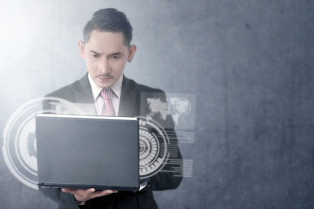 Attractive asian businessman looking at the laptop with virtual screen which display the interface of world map and binary code over wall background. Digital technology conceptの写真素材