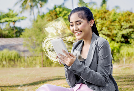 Beautiful asian business woman using digital tablet on her hand with virtual screen which display the interface of world map and binary code on the park. Digital technology conceptの写真素材