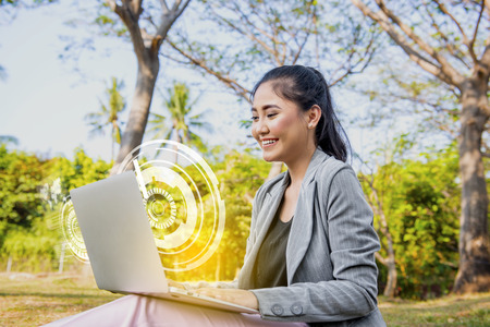 Smiling asian business woman looking at the laptop with virtual screen which display the interface of world maps and binary code on the park. Digital technology conceptの写真素材