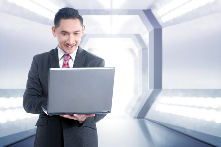 Young asian businessman holding laptop with his hand while standing on the office corridor. Future technology conceptの写真素材