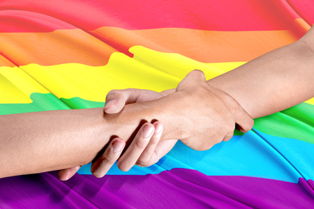 Two gay couple hands holding each other by the wrists with rainbow flag background. LGBT conceptの写真素材