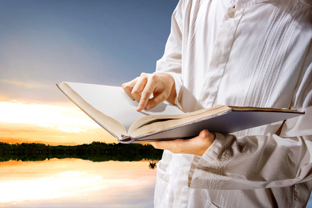 Muslim man standing while reading the quran on his hands at outdoor with lakes and sunset backgroundの写真素材