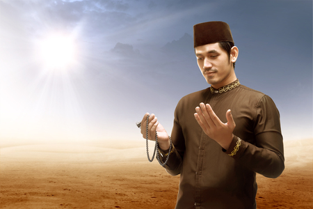Asian muslim man standing and praying while raised arms with prayer beads on desert with sun rays and dark sky backgroundの写真素材