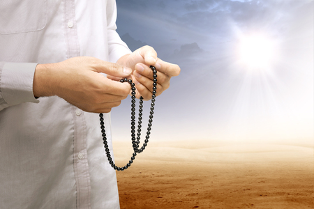 Muslim man praying with prayer beads on his hands on desert with sun rays and dark sky backgroundの写真素材