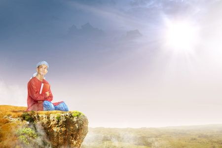 Asian muslim man with turban in his head sitting and holding the quran on the edge of the cliff with sunlight and landscapes backgroundの写真素材