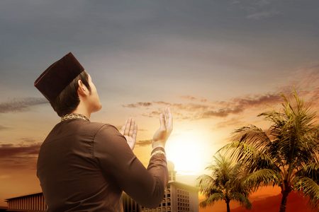 Rear view of asian muslim man standing and praying while raised hands with beautiful mosque with palm trees and sunset on the sky backgroundの写真素材