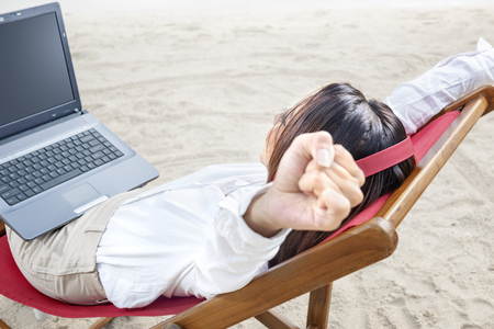 Rear view of asian business woman relax when working with laptop while using headphones sitting in the beach chair on beach. Summer vacationの写真素材