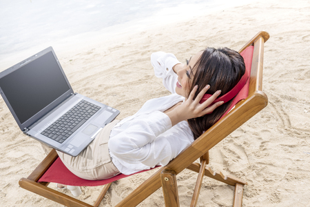 Rear view of asian business woman working with laptop while using headphones sitting in the beach chair on beach. Summer vacationの写真素材