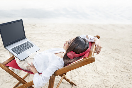 Rear view of asian business woman relax when working with laptop while using headphones sitting in the beach chair on beach. Summer vacationの写真素材