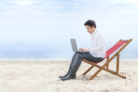 Asian businessman working with laptop sitting in the beach chair on beach. Summer vacationの写真素材
