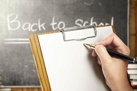Student hands holding clipboard and writing in the classroom with piles of books and blackboard background. Back to School conceptの写真素材